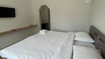 A bedroom with a wooden bed frame, white bedding, a mounted TV, and a small shelf.