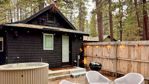 A cabin with a dark exterior, a small porch, a hot tub, and a wooden fence. - The Place Beyond The Pines - Two Bedroom  / C6 (Big Bear Lake)