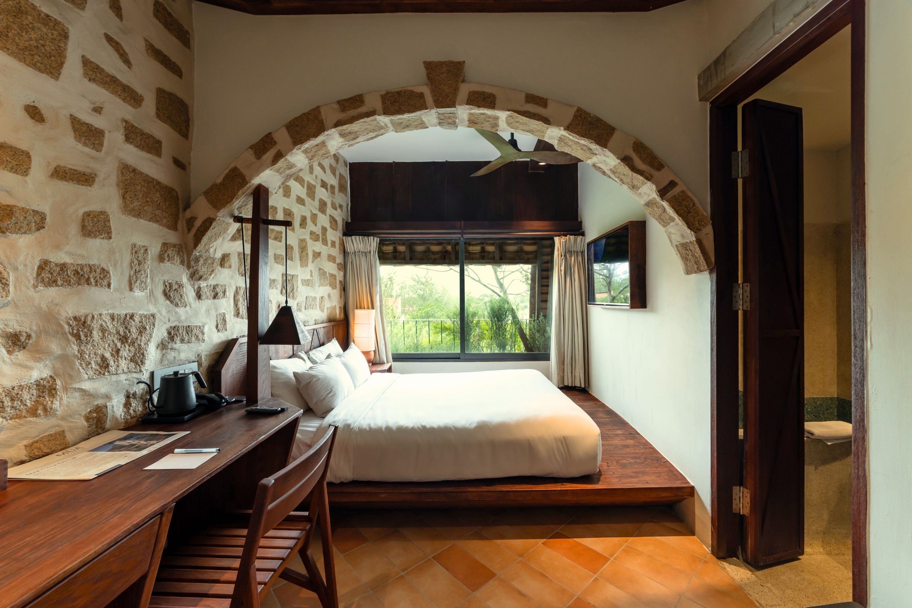 Un dormitorio con pared de piedra, cama, escritorio y ventana con cortinas.