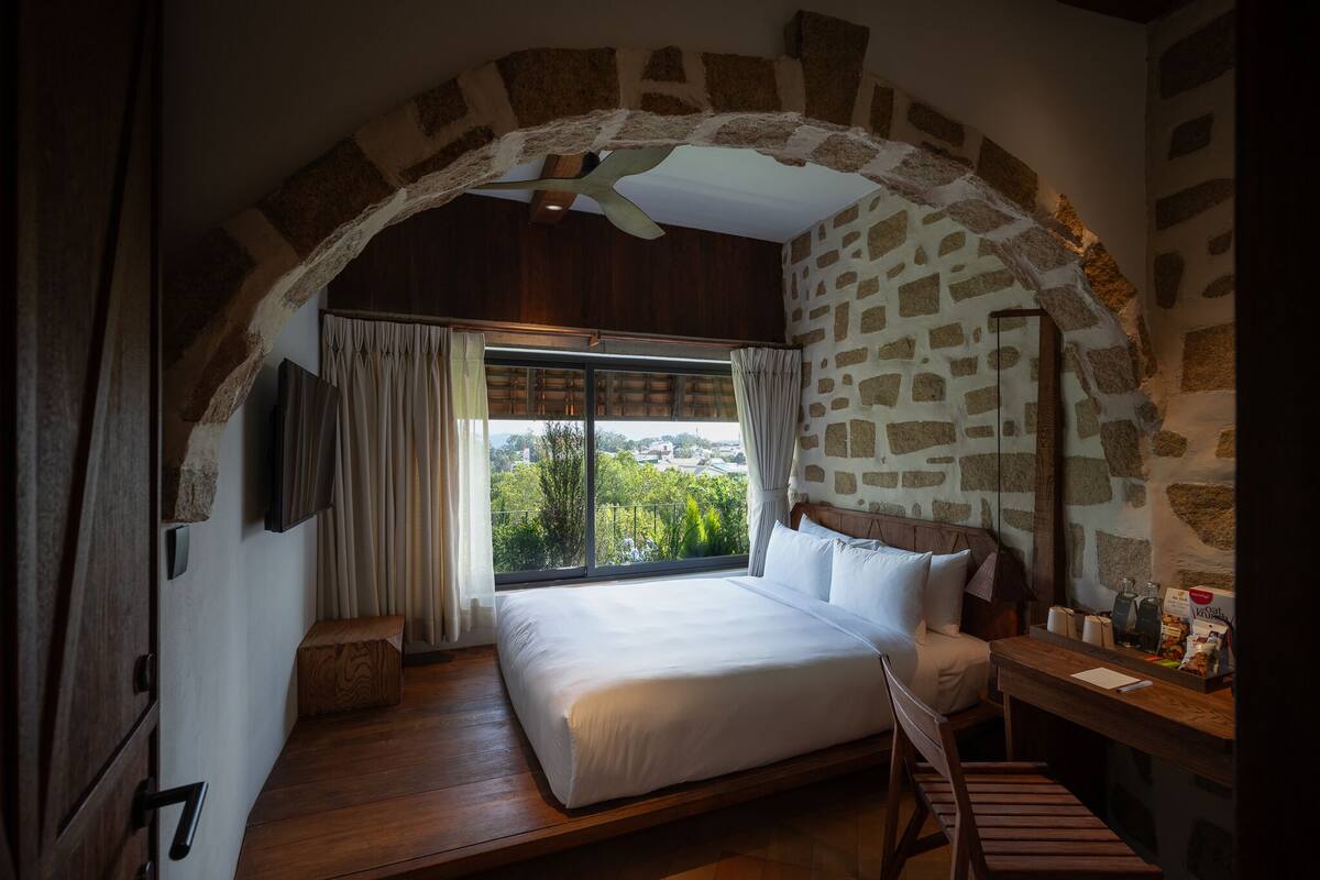 A bedroom with a stone arch ceiling, a large bed, a wooden desk, and a view of greenery outside.