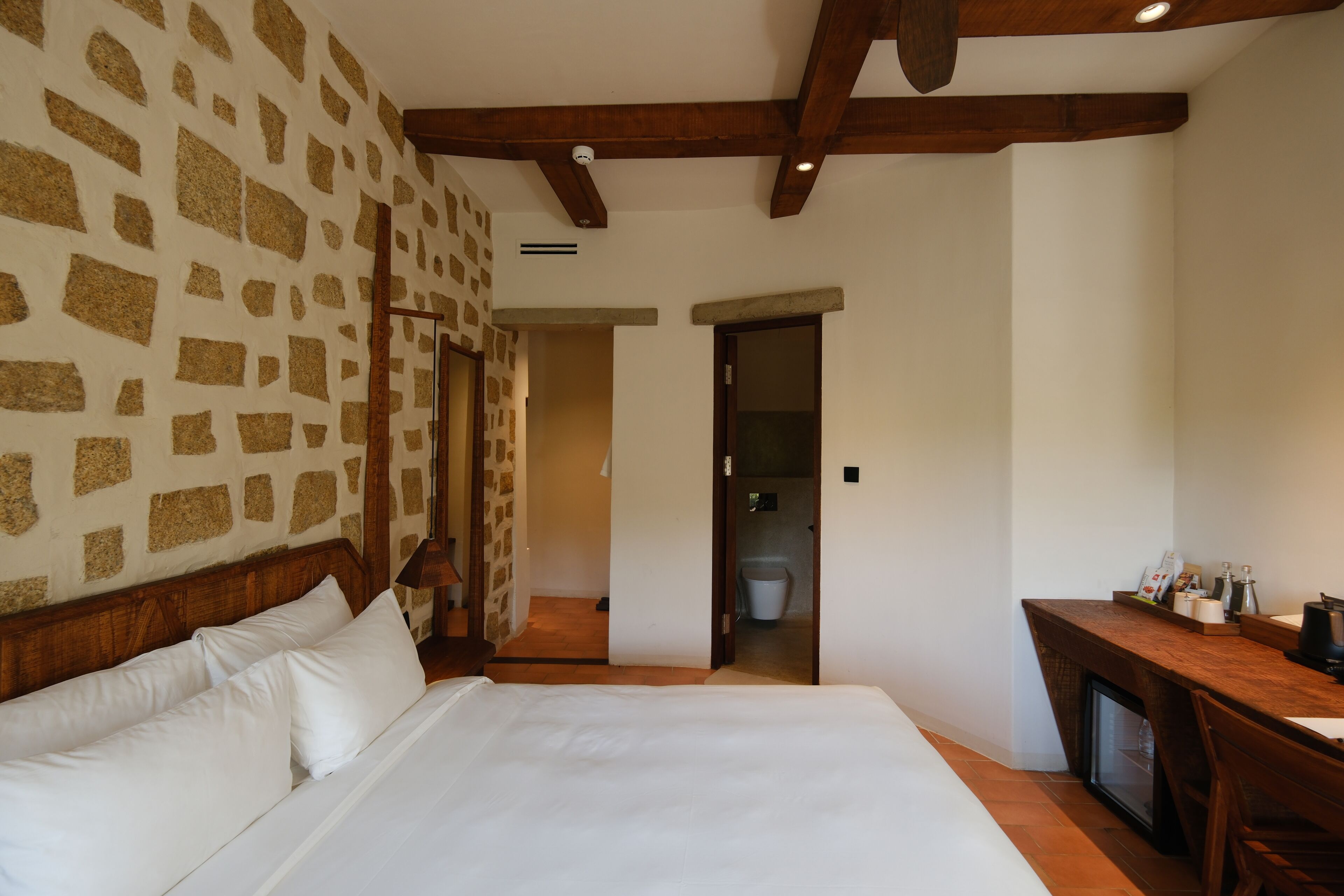 Une chambre avec un mur décoré d’un motif en pierre, une tête de lit en bois, un lit avec du linge de maison blanc, un bureau avec un ordinateur et une porte donnant sur une salle de bain.