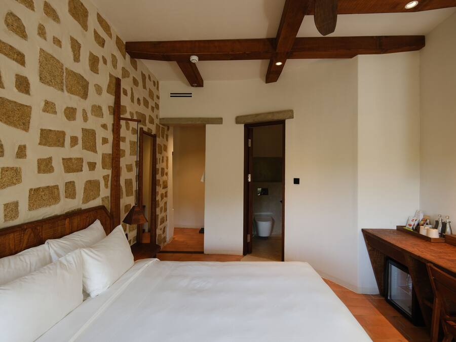 Une chambre avec un mur décoré d’un motif en pierre, une tête de lit en bois, un lit avec du linge de maison blanc, un bureau avec un ordinateur et une porte donnant sur une salle de bain.