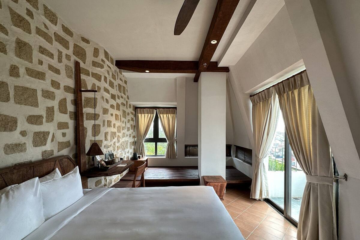 A bedroom with a stone wall, a large bed, a desk, and a window with curtains.