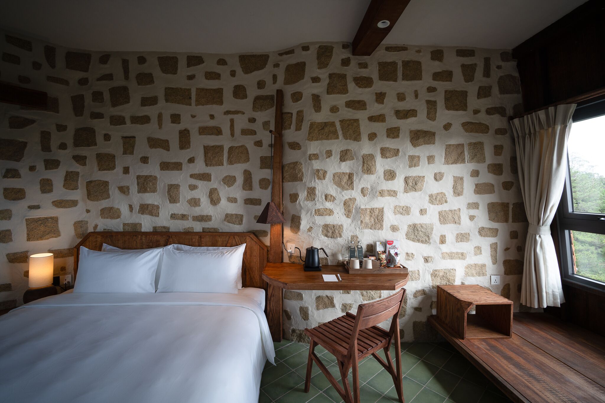 A bedroom with a stone-patterned wall, a wooden bed, a bedside table with a lamp, a small wooden table, and a chair.