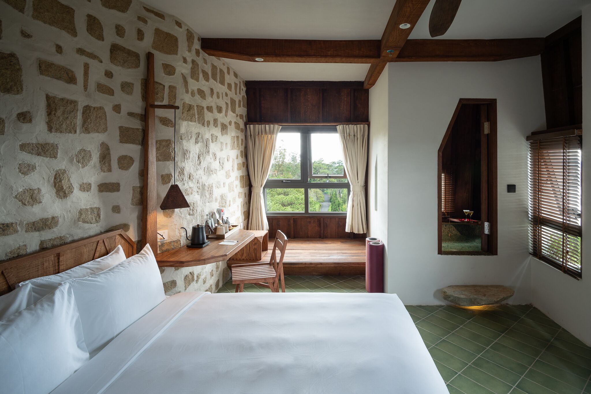 A bedroom with a stone wall, wooden beams, a bed, a desk, and a window with curtains.