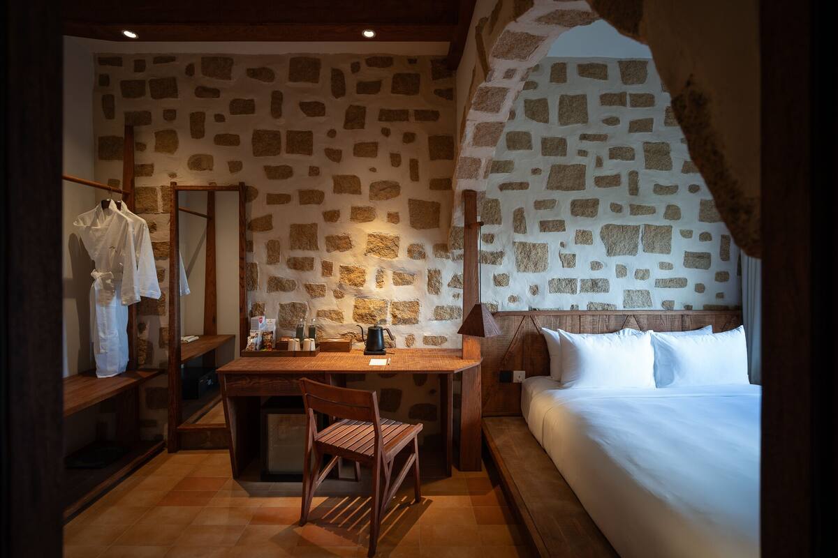 A cozy room with a stone wall, a bed, a desk with a chair, and a hanging robe.