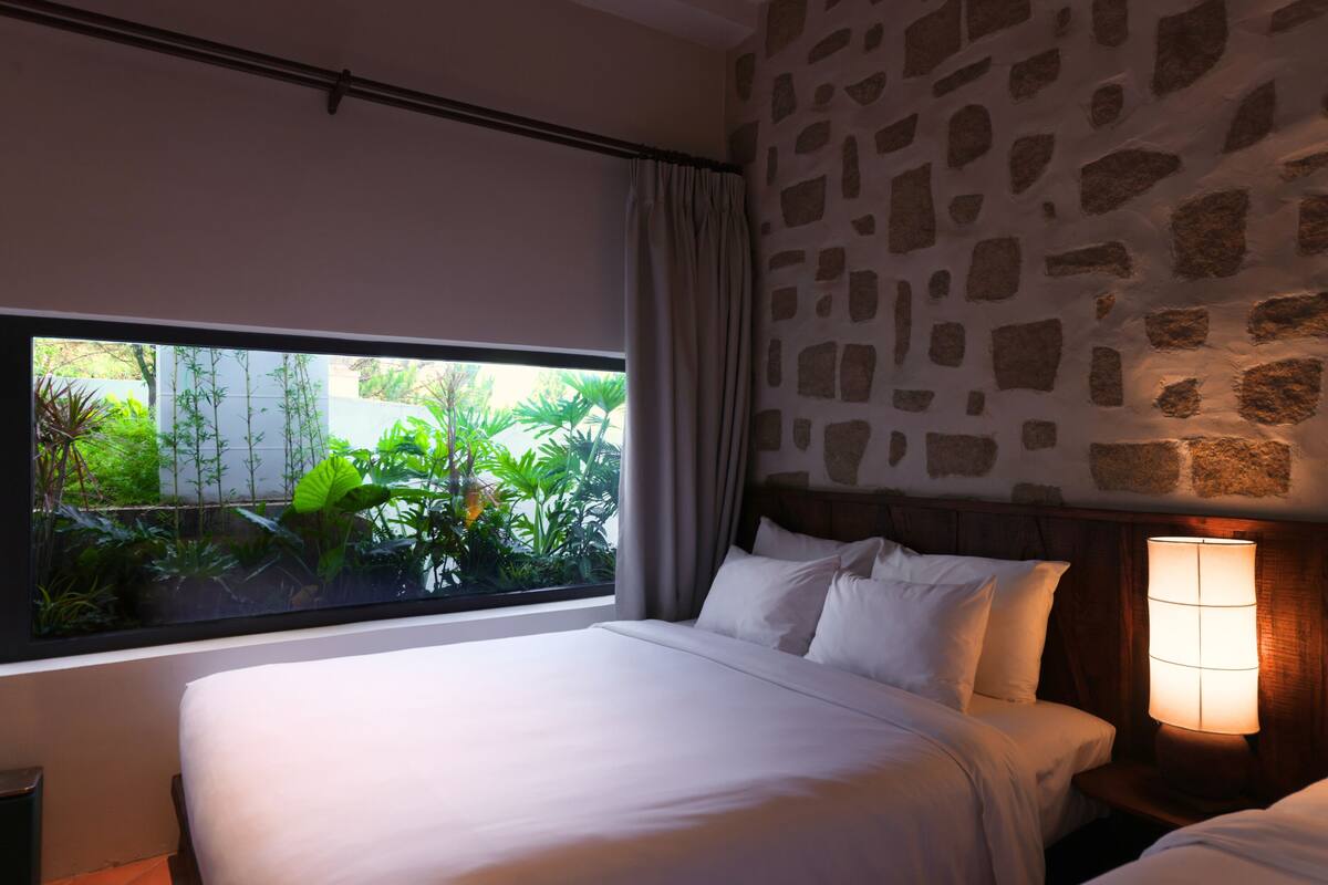 A bedroom with a large window, a stone wall, and a bedside lamp.