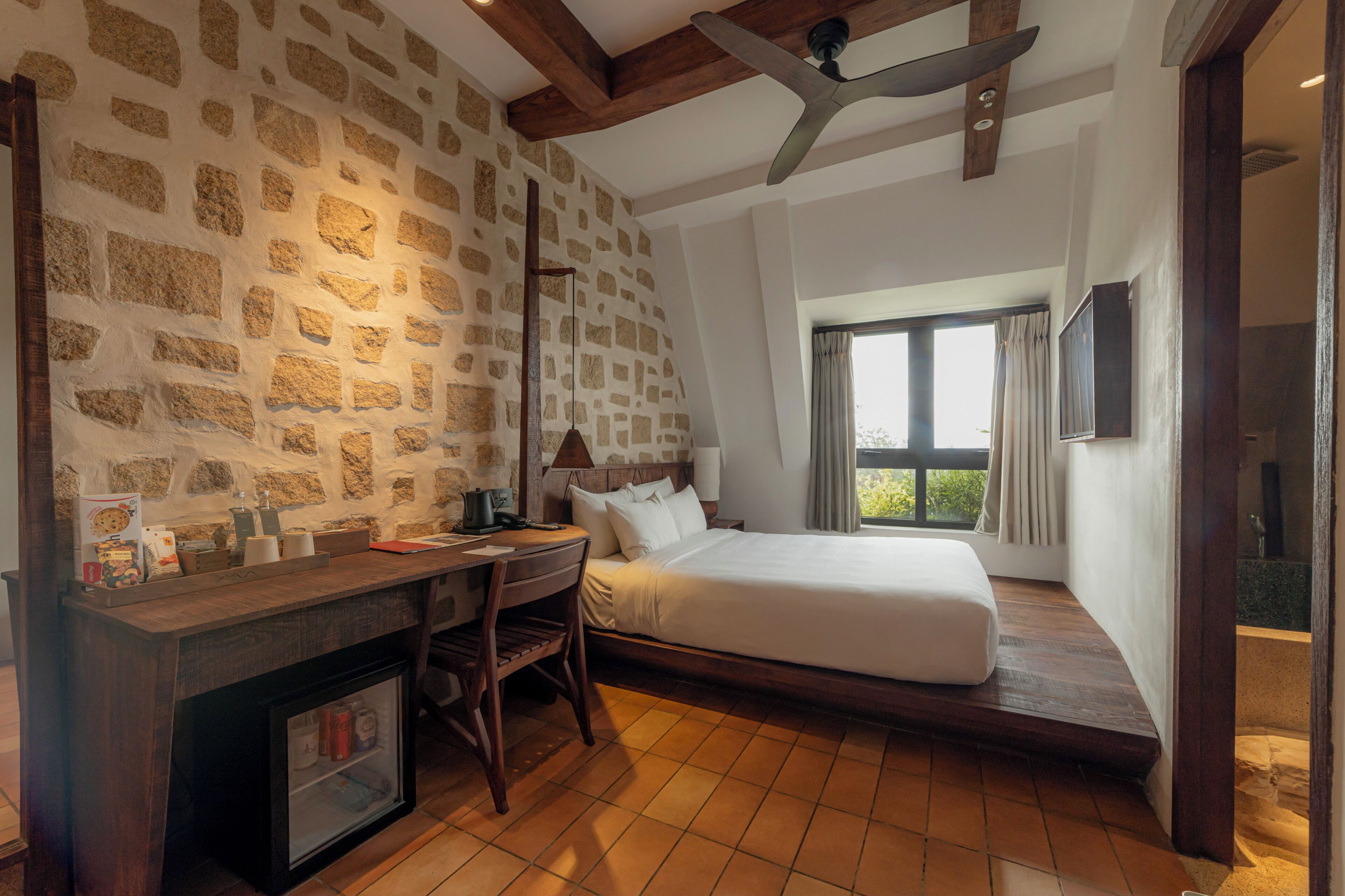 A bedroom with a stone wall, a bed, a desk with a computer, and a window with a view.