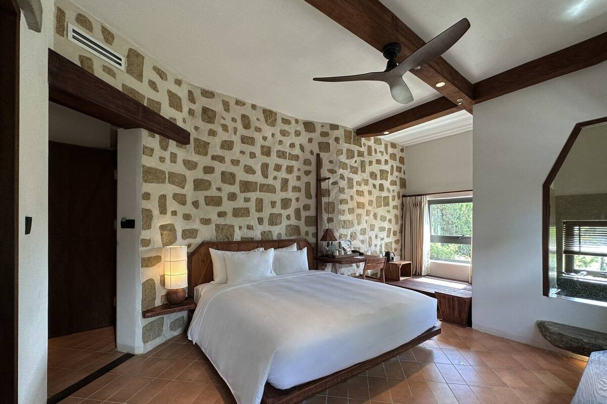 A bedroom with a stone-patterned wall, a large bed, a ceiling fan, and a window with a view of greenery.