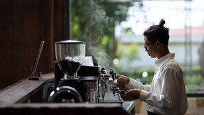 Una persona usando una máquina de café en una habitación con un ventanal grande, paredes de madera y vistas a espacios verdes.