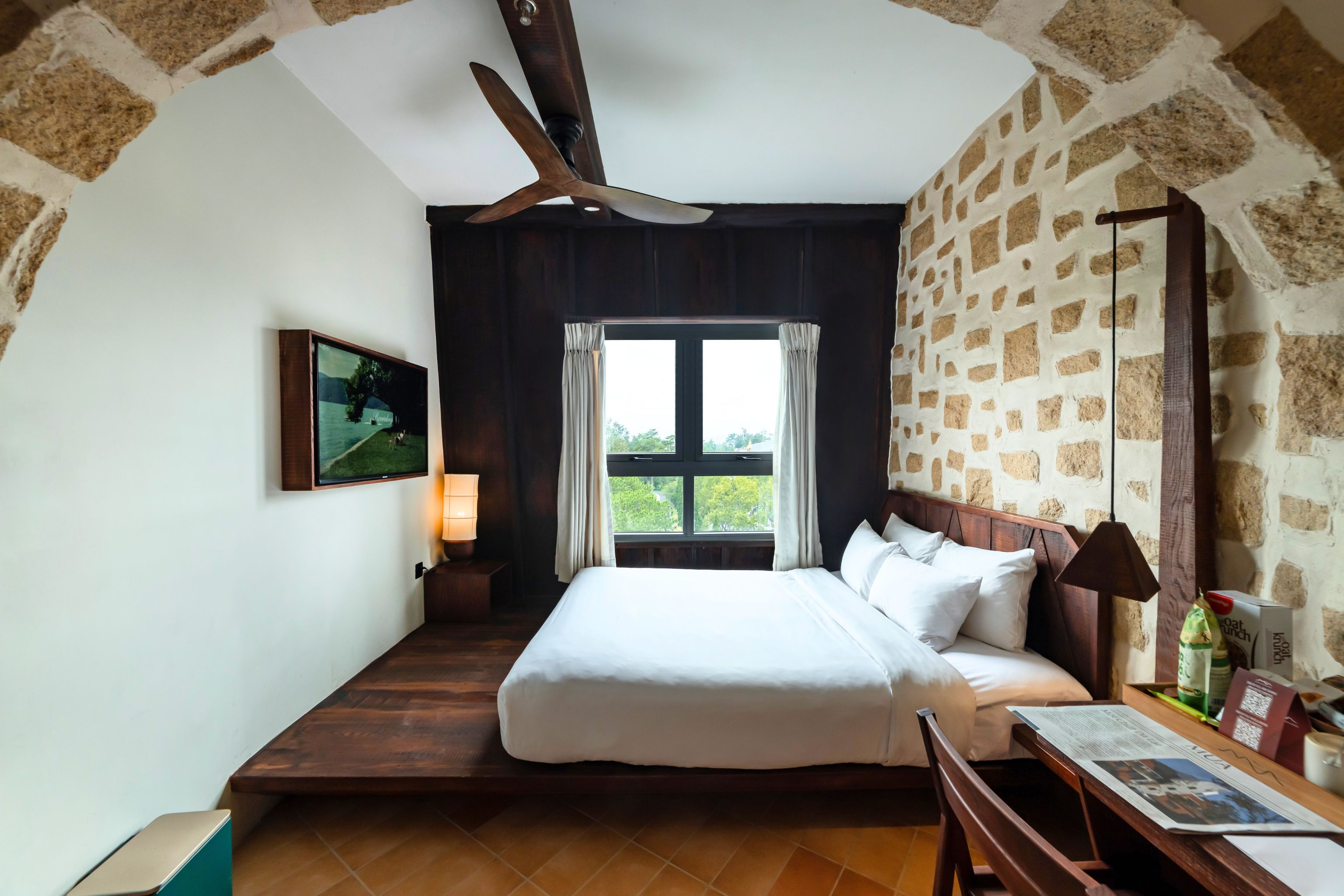 A bedroom with a bed, a ceiling fan, a window with curtains, a wall-mounted TV, and a bedside table with a lamp.