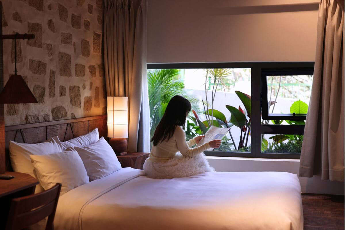 A room with a bed, a nightstand, a lamp, a window with curtains, and a view of greenery outside.