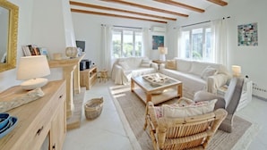 A spacious living room with a high ceiling, wooden beams, a large window, and a cozy seating area with a sofa and a wicker chair. - Vacation home Aika by Interhome (Jávea)