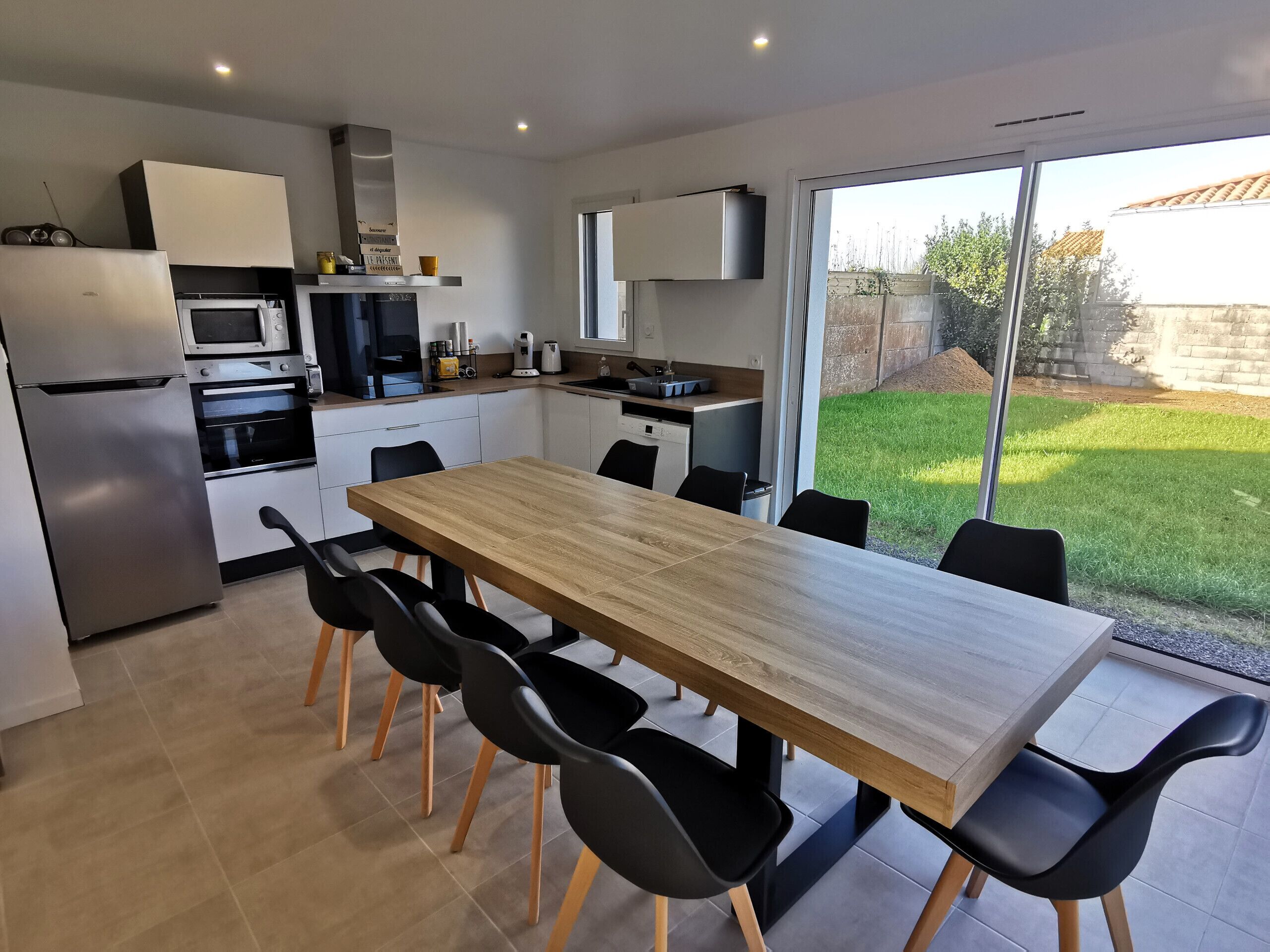 A modern kitchen with a dining area, featuring a large wooden table and black chairs, a built-in oven, and a refrigerator.