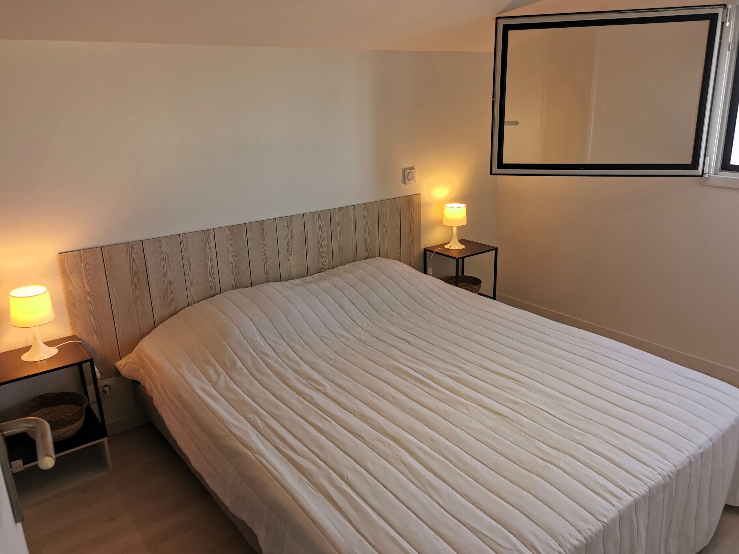 A neatly made bed with a wooden headboard, two bedside lamps, and a small bedside table with a basket.
