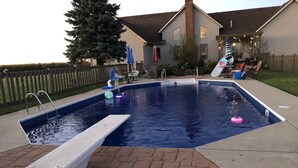 A rectangular swimming pool with a diving board, surrounded by a brick patio and a wooden fence.
