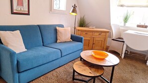 Ein blaues Sofa mit zwei Kissen, ein kleiner runder Tisch mit einer orangen Schale, ein Holzschrank und eine Lampe.