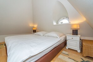 Une chambre à coucher avec un lit, des tables de chevet, une lampe, un panier en osier et une petite fenêtre décorée de fleurs.