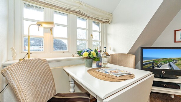 Ein gemütliches Zimmer mit einem weißen Tisch, gestreifte Stühle, einer Lampe, einem Fernseher und einem Fenster mit Vorhängen.