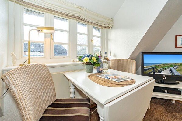 A cozy room with a white table, striped chairs, a lamp, a TV, and a window with curtains.