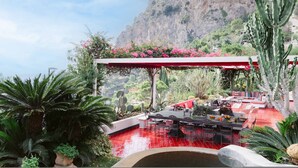 Un patio con piso rojo, rodeado de cactus y palmeras, con vista a un paisaje montañoso.