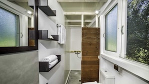 Une petite salle de bain comprenant des toilettes, une douche et un lavabo. On y trouve des étagères en bois avec des serviettes et un miroir au-dessus du lavabo.
