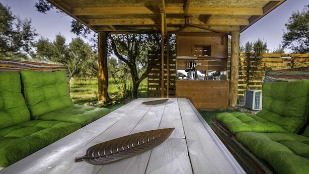 Une structure rustique en bois, dotée de sièges rembourrés de tissu vert et d’une table, le tout entouré d’arbres.