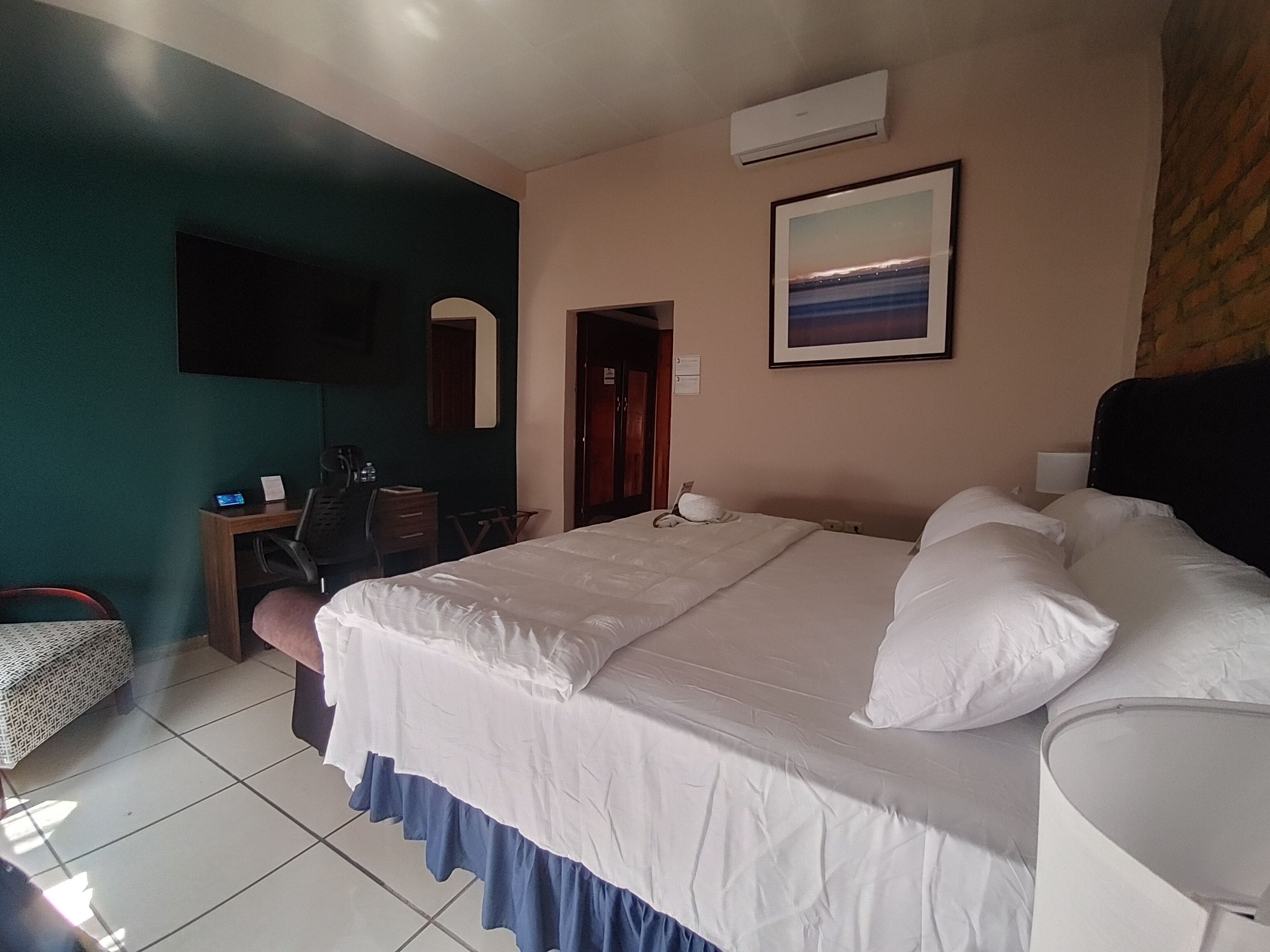 Une chambre d’hôtel avec un grand lit, un bureau avec une chaise, un téléviseur fixé au mur et un tableau encadré au-dessus du lit.