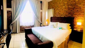 A hotel room with a large bed, a wooden headboard, a brick wall, a window with curtains, a bedside table with a lamp, and a small bench. - Hotel el Campanario (Comayagua)