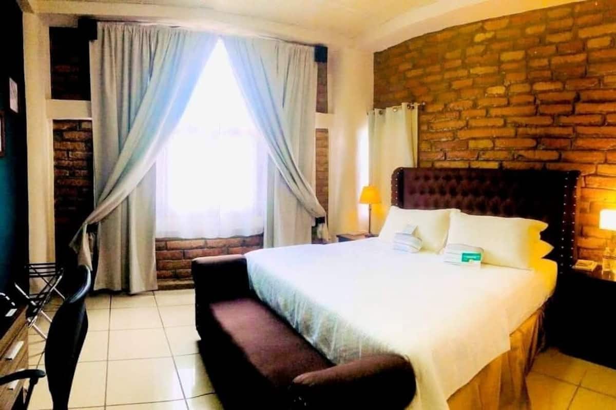 Ein Hotelzimmer mit einem großen Bett, einem Holzkopfteil, einer Ziegelwand, einem Fenster mit Vorhängen, einem Nachttisch mit Lampe und einer kleinen Bank.