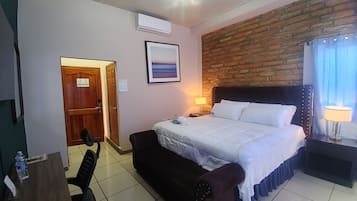 Een hotelkamer met een bed, een bureau met stoel, een houten deur, een bakstenen muur en een raam met gordijnen.