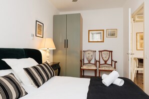 Una camera d'albergo con un letto, un comodino, una lampada, un armadio, due sedie e una porta che conduce a un'altra stanza.