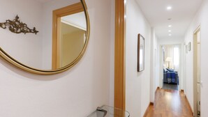 A hallway with a wooden floor, a glass shelf, a round mirror, and a door leading to a room with a sofa and a lamp.