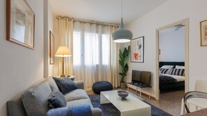 A modern living room with a sofa, coffee table, and a TV. There is a plant, a lamp, and a bed in the background.
