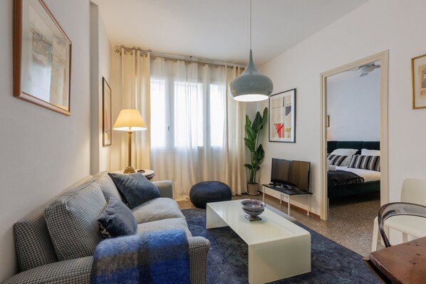 Un soggiorno moderno con divano, tavolino e televisore. Si nota anche una pianta, una lampada e un letto sullo sfondo.
