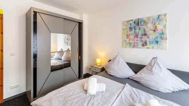 Une chambre d’hôtel avec un lit, une armoire, une table de chevet avec une lampe, et une peinture abstraite aux couleurs vives accrochée au mur.