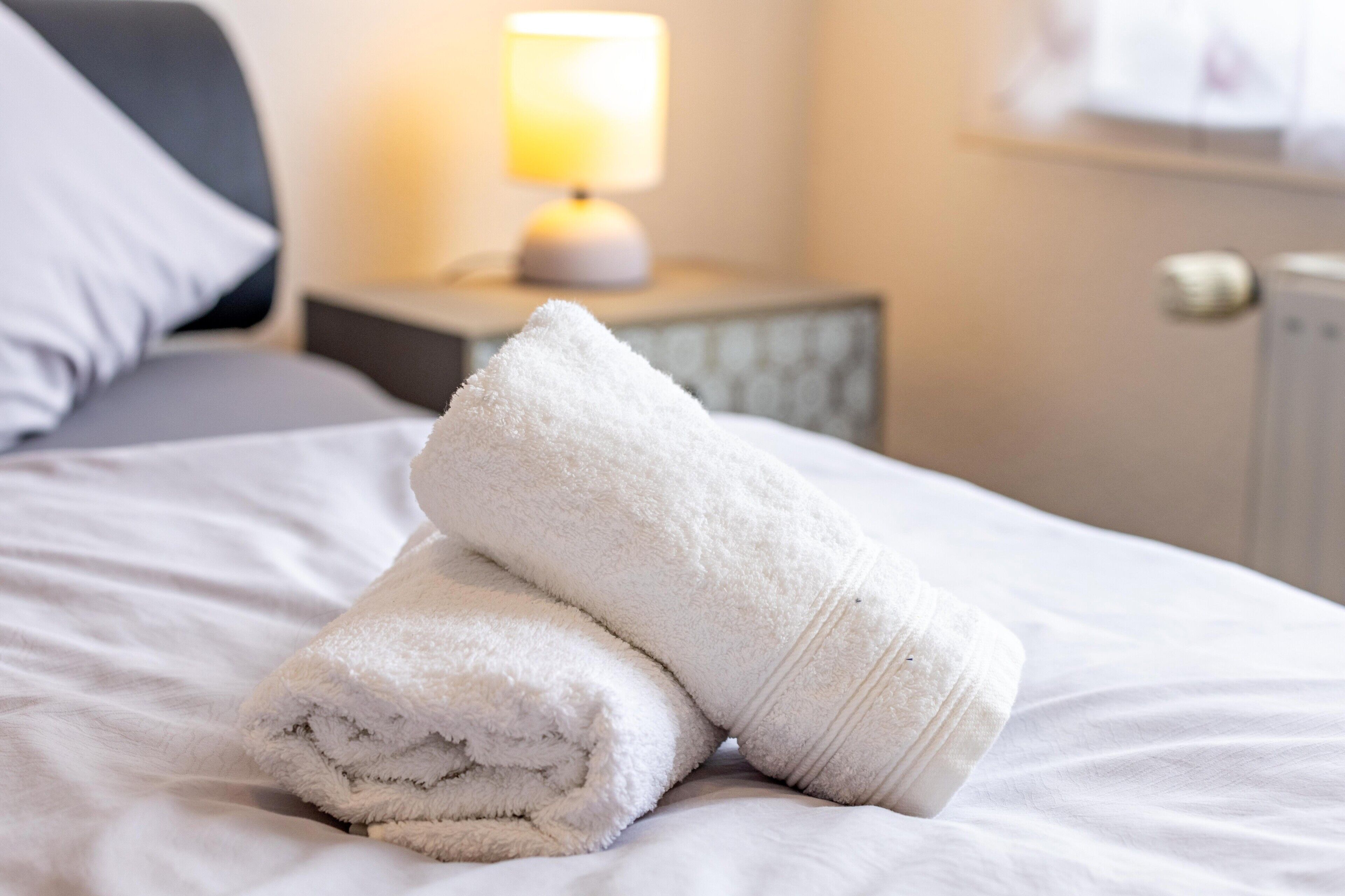 Cama bem arrumada com toalhas brancas dobradas, uma luminária de cabeceira e uma mesa de apoio.