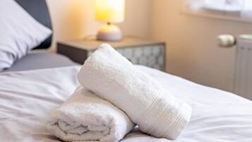 Cama bem arrumada com toalhas brancas dobradas, uma luminária de cabeceira e uma mesa de apoio.