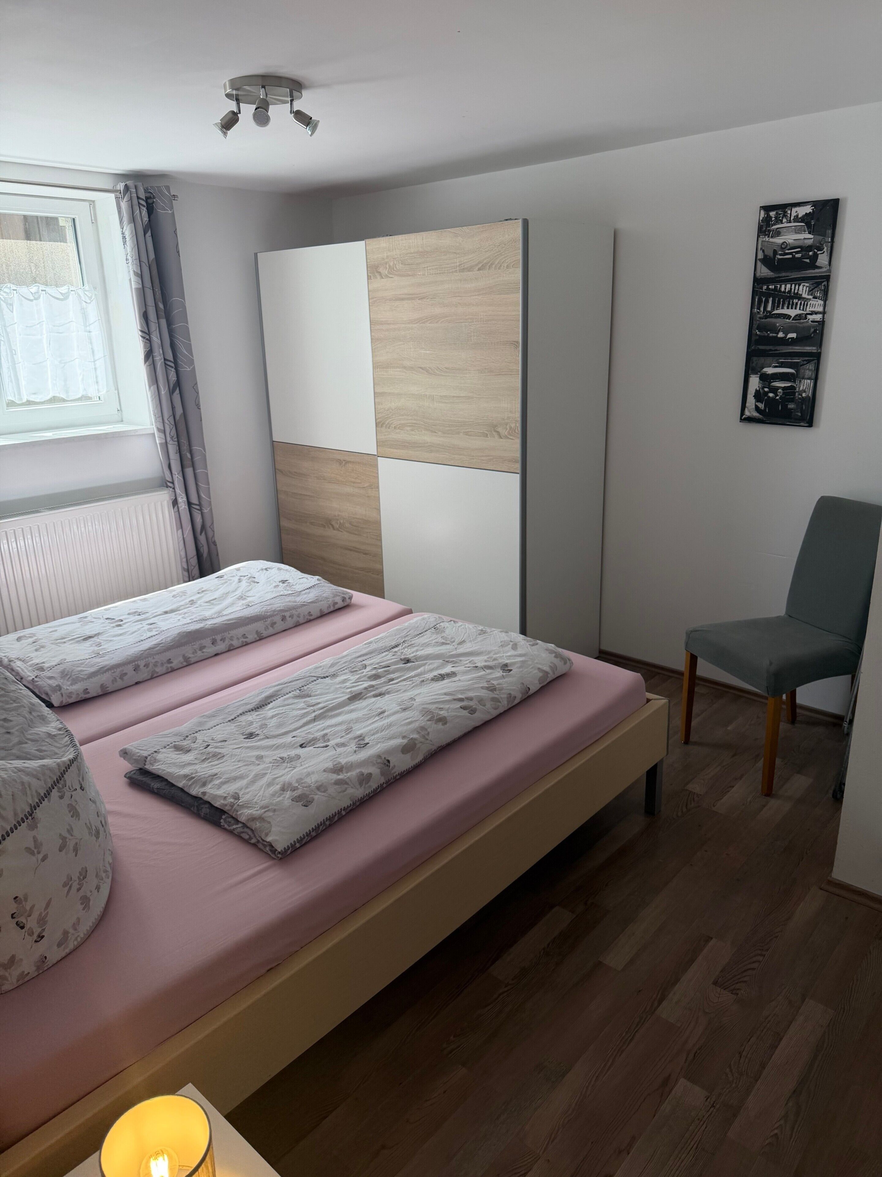 A bedroom with a bed, a chair, a window with curtains, a wall-mounted light fixture, and a framed picture.