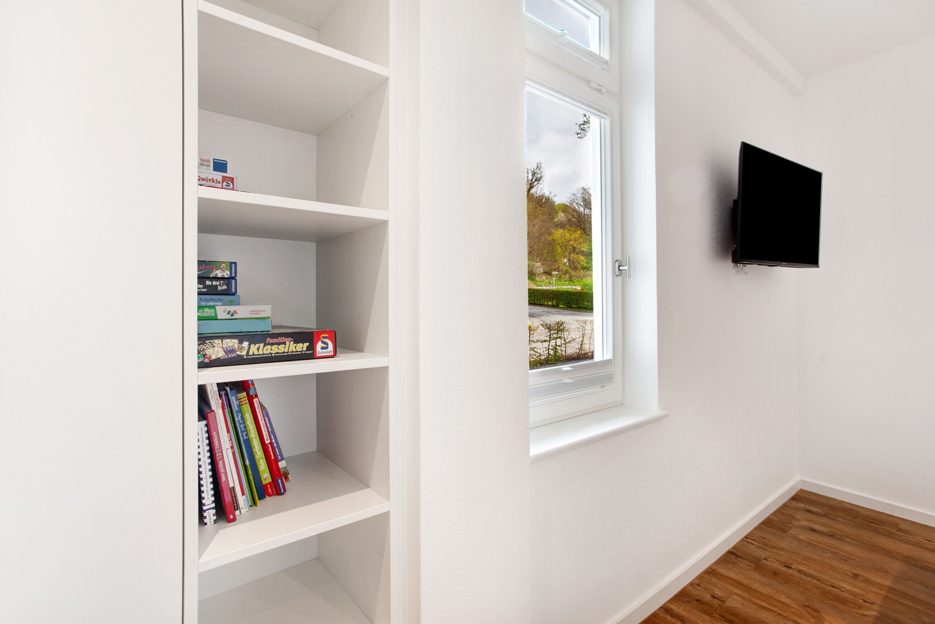 A room with white walls, a bookshelf with books, a window with a view, and a mounted TV.