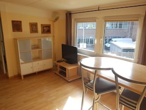 Ein Zimmer mit Holzboden, einem Fernsehgerät auf einem Standfuß, einem Esstisch mit Stühlen und einem Fenster mit Blick auf Gebäude.