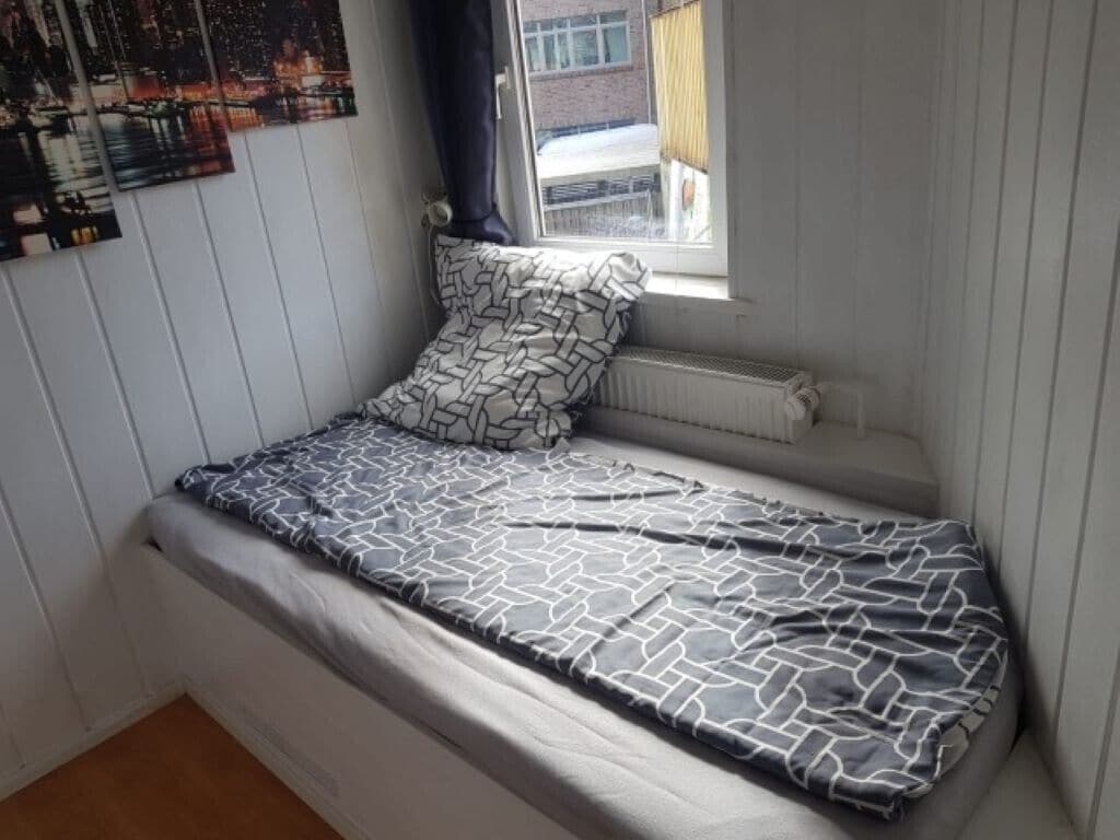 A small, single-bed room with a window, a radiator, and a wall-mounted artwork.