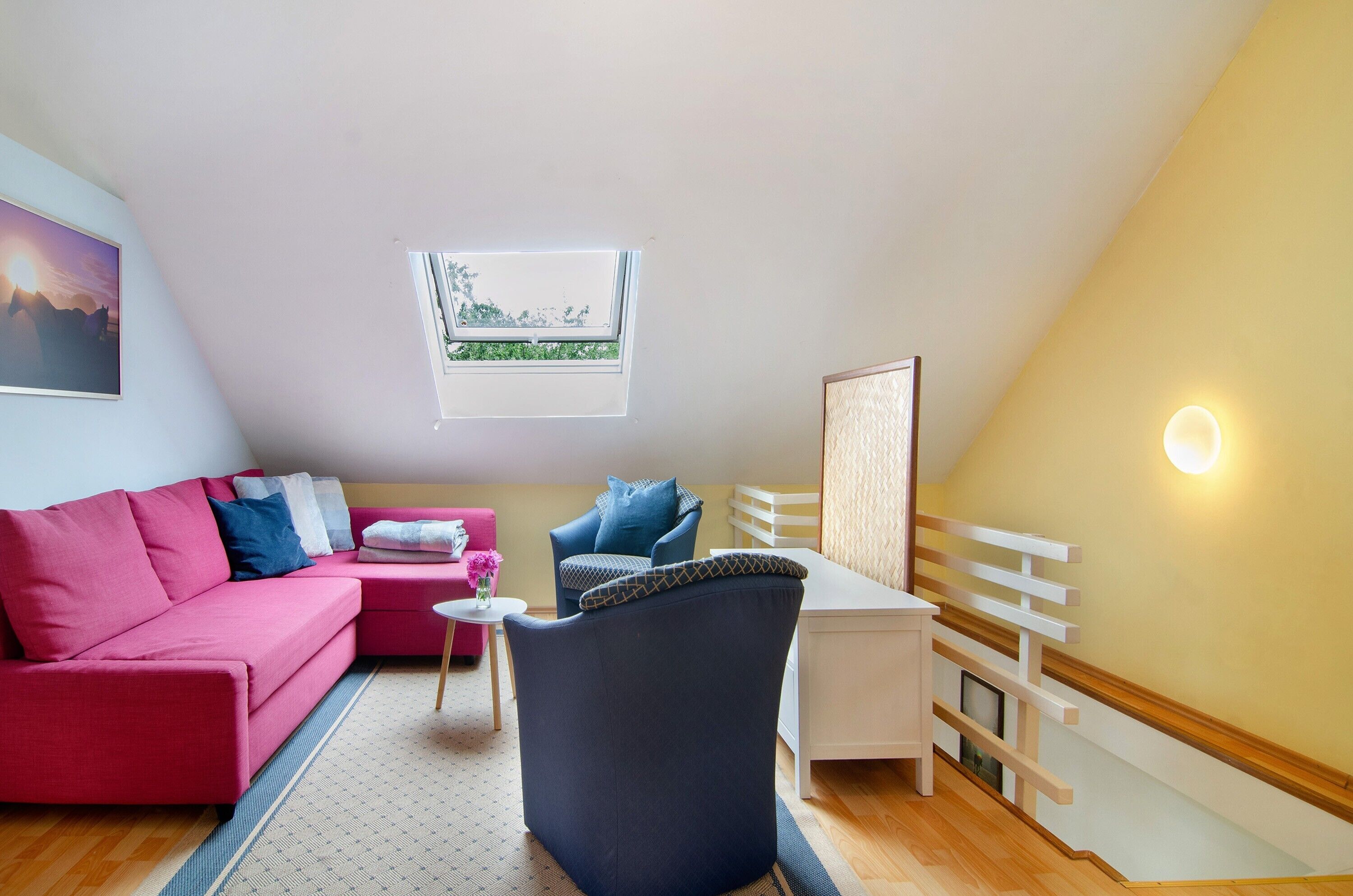 A cozy room with a sloped ceiling, a red sofa, a blue armchair, a small white table, and a window with a view of greenery.