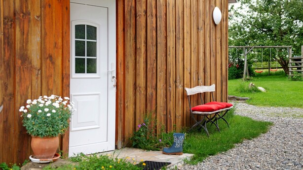 Un bâtiment en bois avec une porte blanche, un petit espace extérieur aménagé avec un coussin rouge et une plante en pot.