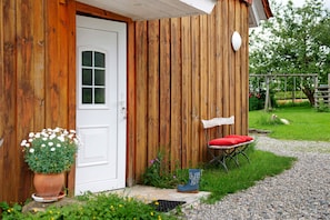 A wooden building with a white door, a small outdoor seating area with a red cushion, and a potted plant.