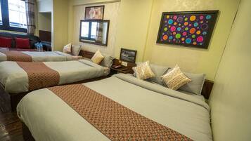 A hotel room with three beds, each with a patterned bedspread and pillows. There are two framed pictures on the wall, one with a floral design and another with a colorful abstract pattern.