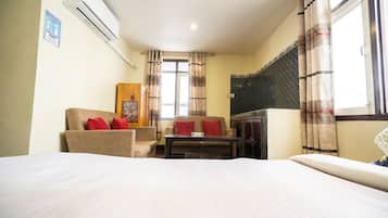 A hotel room with a bed, a sofa, a small table, and a cabinet. There is a window with curtains, an air conditioning unit on the wall, and a poster on the wall.