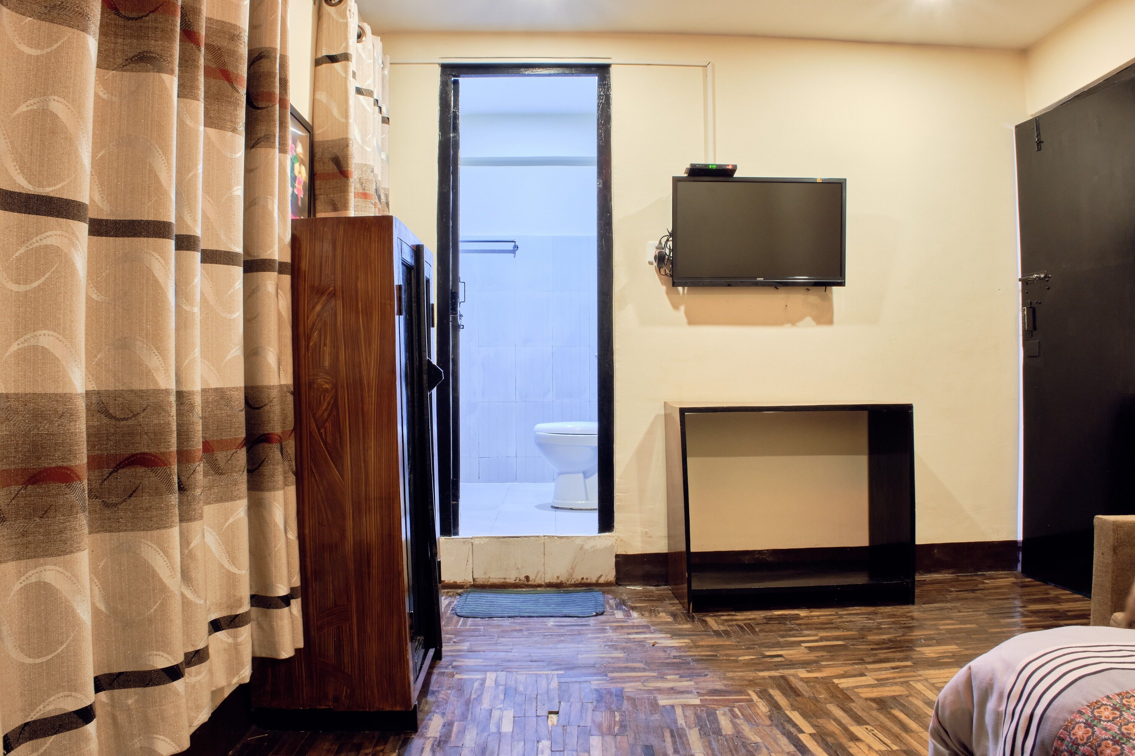 A hotel room with a wooden wardrobe, a television mounted on the wall, a small table, and a door leading to a bathroom.