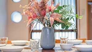 Una mesa de comedor dispuesta con platos blancos, tazones y una copa de cristal transparente. Un florero con follaje rosa y verde está centrado en la mesa.