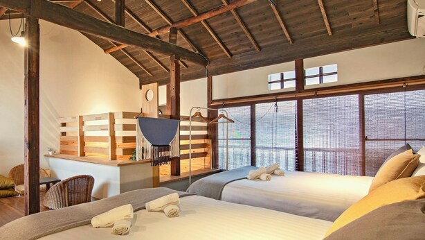 Una habitación con dos camas, techo de madera, un escritorio pequeño y ventanas amplias.